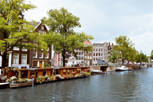 Ansicht von einer Gracht in Amsterdam mit traditionellen Häusern und Hausbooten bei bewölktem Himmel. Am Ufer sind Bäume, Fahrräder und Passanten.