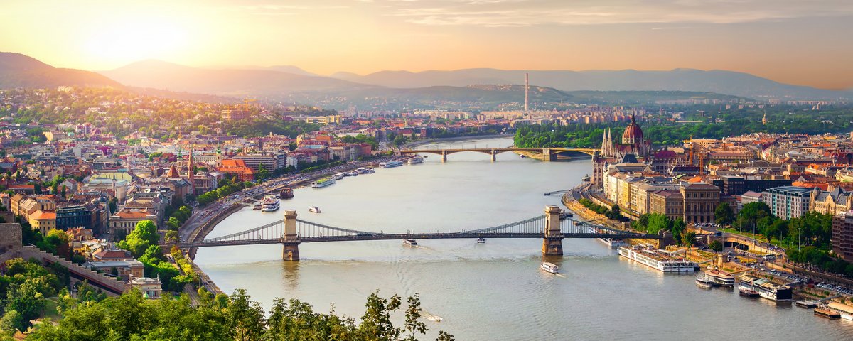 Panoramablick auf Budapest mit der Kettenbrücke über die Donau, im Hintergrund das Parlamentsgebäude und die Budaer Burg bei Sonnenuntergang.