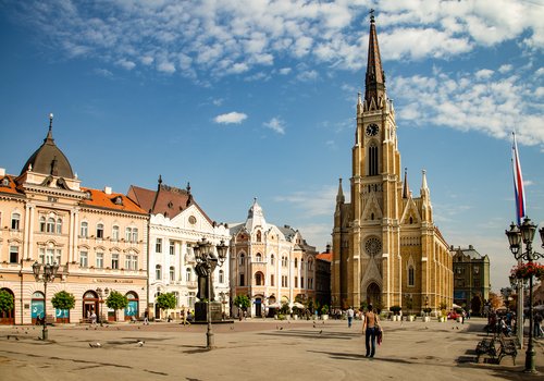 Blick auf einen Marktplatz in der Statd Novi Sad mit eier bräunlichen Kirche im Hintergrund umgeben von historischen Gebäuden.