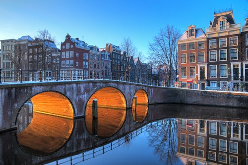 Brücke mit drei Bögen über einen Kanal in Amsterdam, beleuchtet von warmem Licht, mit historischen Häusern und klarer Spiegelung im Wasser