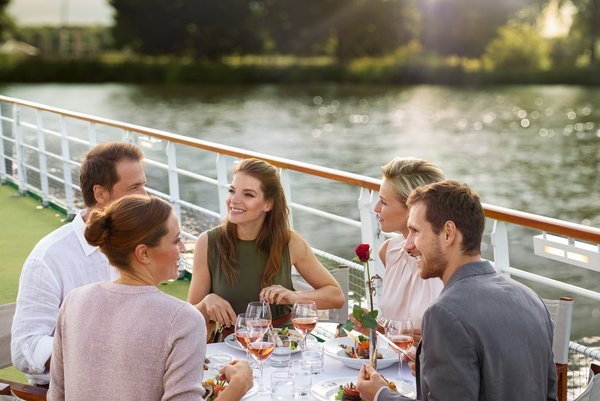 Yvonne Catterfeld und vier weitere Personen sitzen an einem gedeckten Tisch auf einem Sonnendeck am Wasser und genießen ein Essen bei Sonnenuntergang.