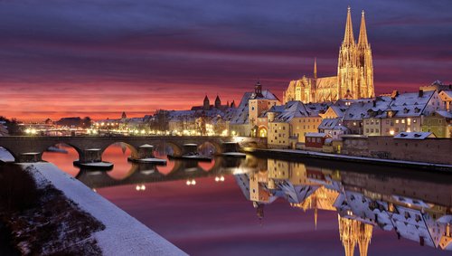 Winterliche Stadtansicht von Regensburg mit schneebedeckten Dächern und der Donau im Vordergrund, und dem Dom im Hintergrund bei Abenddämmerung