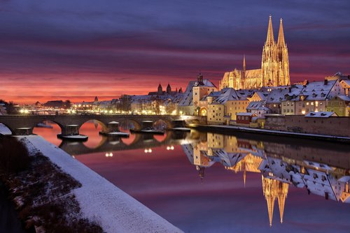 Winterliche Stadtansicht von Regensburg mit schneebedeckten Dächern und der Donau im Vordergrund, und dem Dom im Hintergrund bei Abenddämmerung