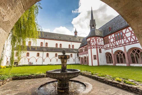 Innenhof des Klosters Eberbach in Mainz mit Brunnen und Wiese, umgeben von Fachwerkarchitektur unter blauem Himmel.