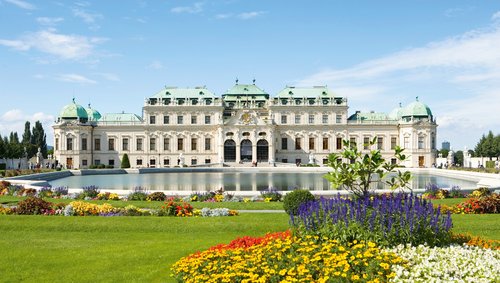 Das Schloss Belvedere in Wien, umgeben von gepflegten Gärten mit bunten Blumen und einem Brunnen bei sonnigem Wetter, mit barocker Architektur und kunstvollen Details.