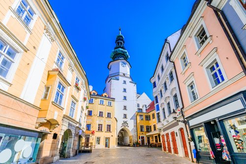 Blick durch eine Gasse der Altstadt von Bratislava mit bunten Häusern am Straßenrand einem weißen Kirchturm im Hintergrund, der Himmel ist wolkenlos und blau.
