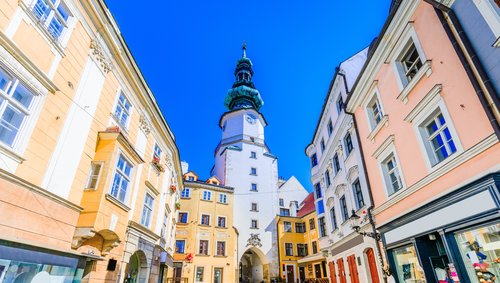 Blick durch eine Gasse der Altstadt von Bratislava mit bunten Häusern am Straßenrand einem weißen Kirchturm im Hintergrund, der Himmel ist wolkenlos und blau.