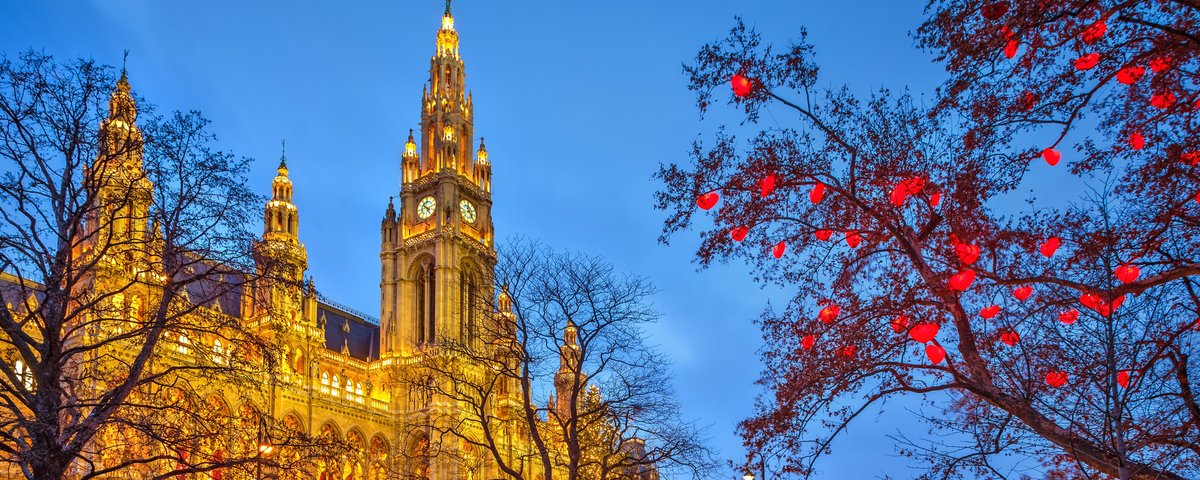 Beleuchtetes Wiener Rathaus bei Dämmerung mit winterlichem Park und roten Lichtern an Bäumen