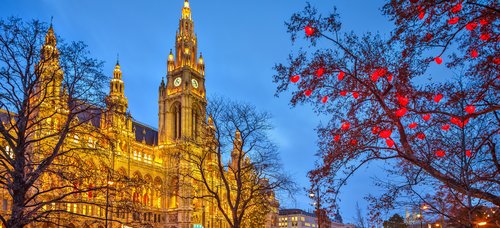 Beleuchtetes Wiener Rathaus bei Dämmerung mit winterlichem Park und roten Lichtern an Bäumen