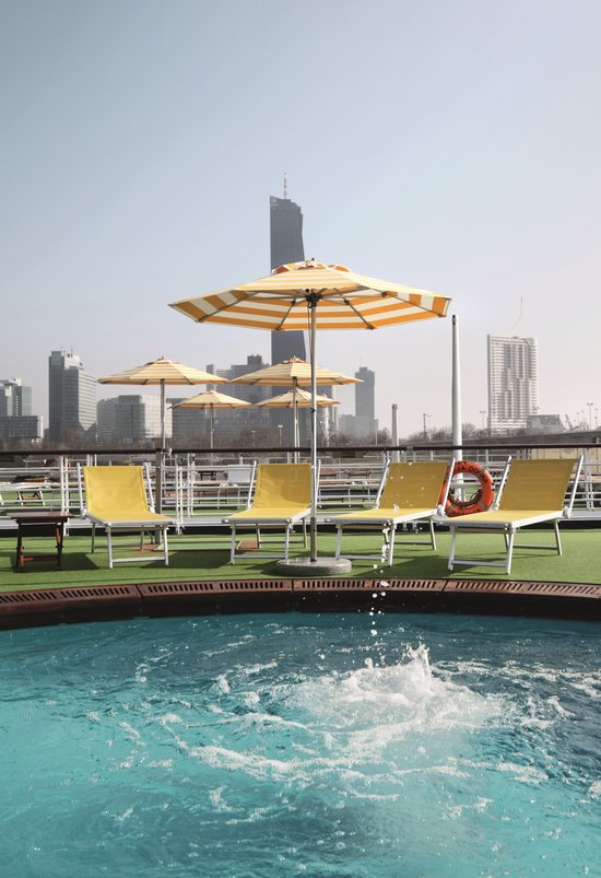 Sonnendeck des Flusskreuzfahrtschiffs A-ROSA DONNA mit Pool im Vordergrund, umgeben von Liegestühlen und Sonnenschirmen, mit Hochhäusern im Hintergrund.