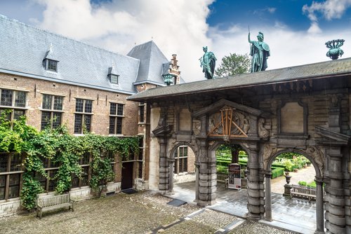Historischer Innenhof in Antwerpen mit einer Steinarkade, die mit drei bronzenen Statuen auf dem Dach verziert ist, umgeben von Gebäuden mit Kletterpflanzen an den Wänden