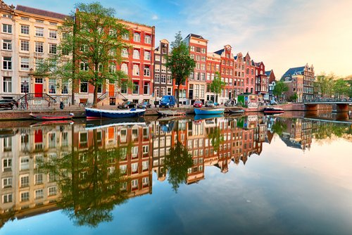 Typische Amsterdamer Grachtenhäuser in verschiedenen Farben spiegeln sich im ruhigen Wasser eines Kanals mit kleinen Booten und Bäumen am Ufer