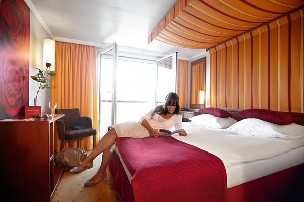 Eine Kabine der Kategorie C und D an Bord der A-ROSA LUNA und A-ROSA STELLA. Einrichtet in warmen Farben mit Doppelbett und einem französischen Balkon. Eine Frau liegt auf dem Bett und liest eine Zeitung.