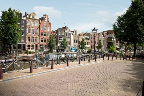 Eine Amsterdamer Gracht mit historischen Gebäuden, Bäumen und Booten. Im Vordergrund ist ein gepflasterer Weg und Fahrräder sind am Zaun am Grachtenufer angebracht.
