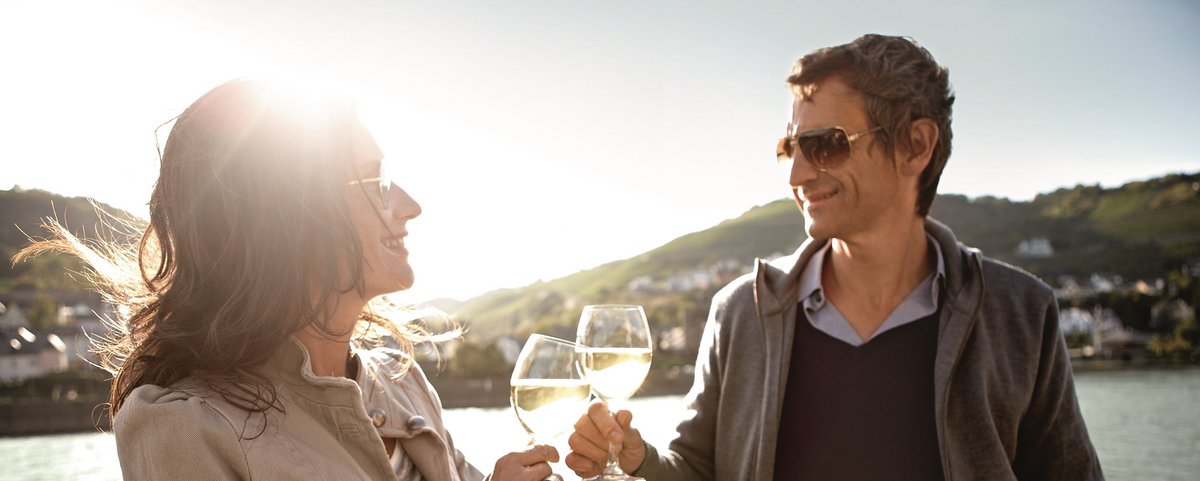 Zwei Personen stehen auf dem Sonnendeck eines Flusskreuzfahrtschiffs und stoßen mit Weißwein an, umgeben von grünen Hügeln mit Häusern und dem Rhein im Hintergrund.