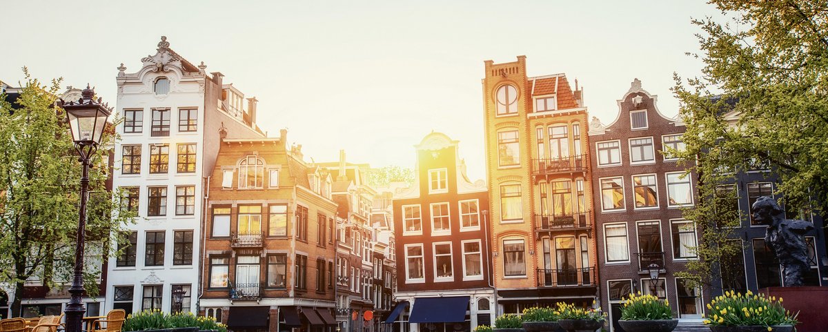 Blick auf Amsterdams historische Gebäude bei Sonnenuntergang. Im Vordergrund ist ein gepflasterter Weg umgeben von Bäumen, Blumentöpfen, einer Laterne und Sitzmöglichkeiten unter klarem Himmel.