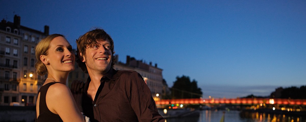 Ein Paar umarmt sich auf einem Flusskreuzfahrtenschiff bei Dämmerung, im Hintergrund eine beleuchteter Brücke und Wasser des Flusses. 