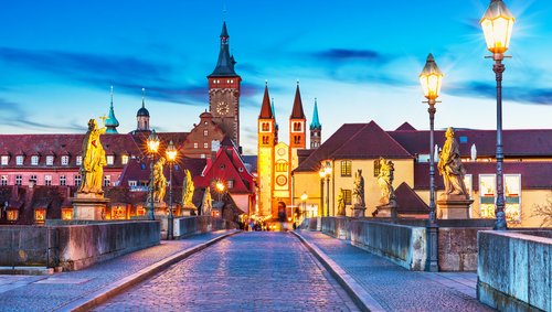 Historische Alte Mainbrücke in Würzburg bei Abenddämmerung mit beleuchteten barocken Statuen und Türmen der Stadt im Hintergrund