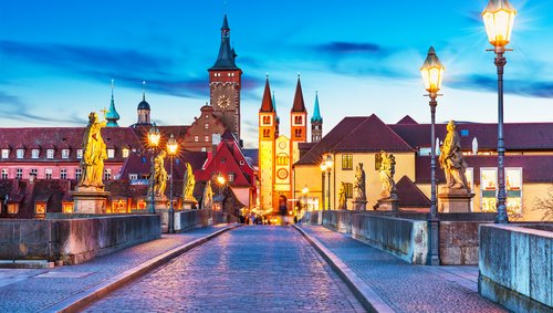 Historische Alte Mainbrücke in Würzburg bei Abenddämmerung mit beleuchteten barocken Statuen und Türmen der Stadt im Hintergrund