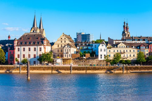 Promenade in Koblenz mit Blick auf den Rhein, gesäumt von Bäumen und historischen Gebäuden, bei klarem Himmel und Sonnenschein.