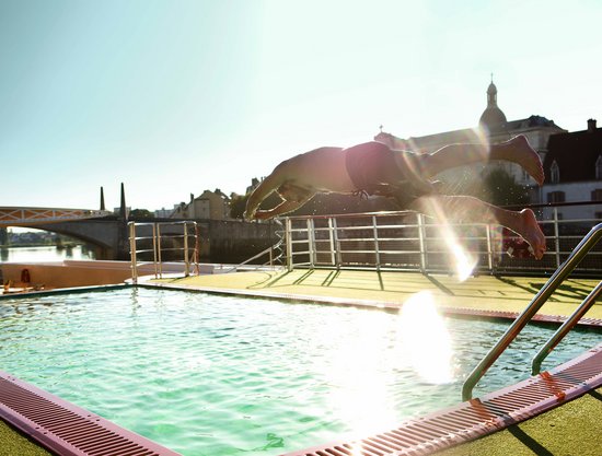 Ein Mann springt vom Beckenrand in den Pool auf dem Sonnendeck eines Flusskreuzfahrtschiffes. Im Hintergrund die Gebäude einer Stadt und die scheinende Sonne.