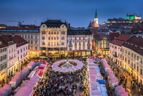 Weihnachtsmarkt in Bratislava am Abend mit beleuchteten Ständen, umgeben von Gebäuden. Im der Ferne liegt Burg Bratislava.