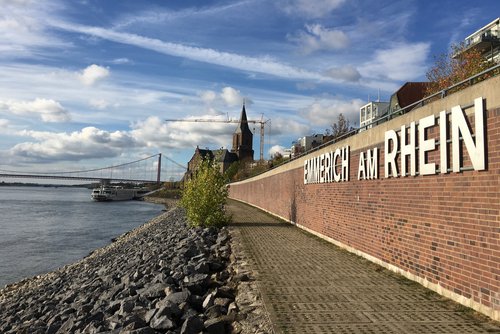 Uferpromenade in Emmerich am Rhein mit Blick auf den Fluss, eine Kirche und eine Hängebrücke unter blauem Himmel mit Wolken.