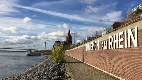 Uferpromenade in Emmerich am Rhein mit Blick auf den Fluss, eine Kirche und eine Hängebrücke unter blauem Himmel mit Wolken.