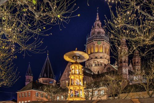 Blick auf den beleuchteten Mainzer Dom bei Nacht, davor die Spitze einer Weihnachtspyramide. Im Vordergrund kahle Bäume, die mit Lichterketten geschmückt sind.