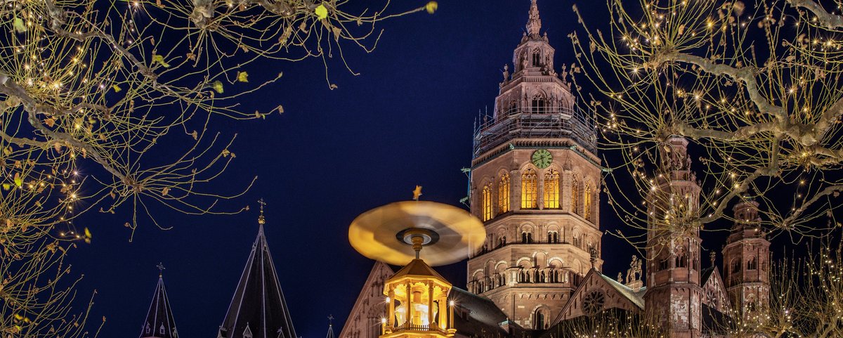 Blick auf den beleuchteten Mainzer Dom bei Nacht, davor die Spitze einer Weihnachtspyramide. Im Vordergrund kahle Bäume, die mit Lichterketten geschmückt sind.
