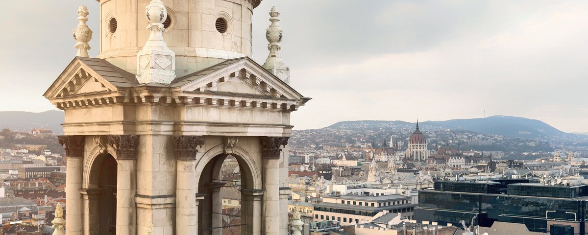 Die St.-Stephans-Basilika in Budapest mit Blick auf die Stadt und das Parlamentsgebäude an der Donau. Im Hintergrund Hügel und ein bewölkter Himmel.