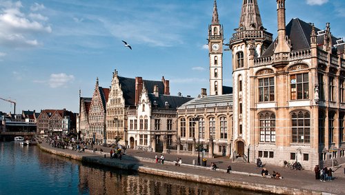 Gent mit historischen Gebäuden am belebten Kanalufer bei Sonnenschein, Boote auf dem Wasser, Menschen spazieren entlang der Uferpromenade.