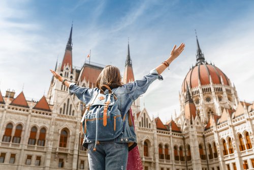 Blick auf eine Frau mit Rucksack von hinten, die ihre Arme nach oben ausstreckt und das ungarische Parlamentsgebäude in Budapest bei sonnigem Wetter bestaunt.