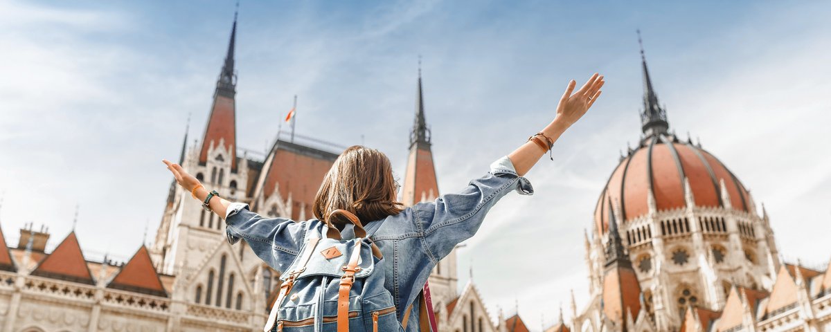Blick auf eine Frau mit Rucksack von hinten, die ihre Arme nach oben ausstreckt und das ungarische Parlamentsgebäude in Budapest bei sonnigem Wetter bestaunt.