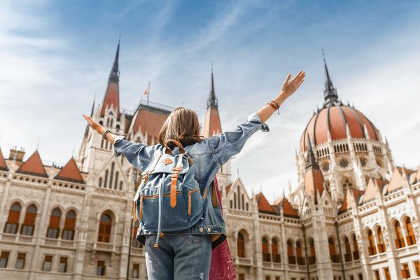 Blick auf eine Frau mit Rucksack von hinten, die ihre Arme nach oben ausstreckt und das ungarische Parlamentsgebäude in Budapest bei sonnigem Wetter bestaunt.
