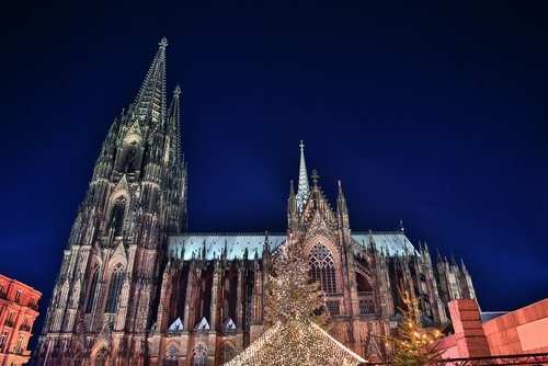 Kölner Dom bei Nacht mit beleuchtetem Weihnachtsmarkt und festlich geschmücktem Weihnachtsbaum im Vordergrund.