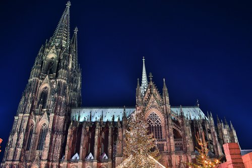 Kölner Dom bei Nacht mit beleuchtetem Weihnachtsmarkt und festlich geschmücktem Weihnachtsbaum im Vordergrund.