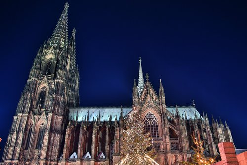 Kölner Dom bei Nacht mit beleuchtetem Weihnachtsmarkt und festlich geschmücktem Weihnachtsbaum im Vordergrund.