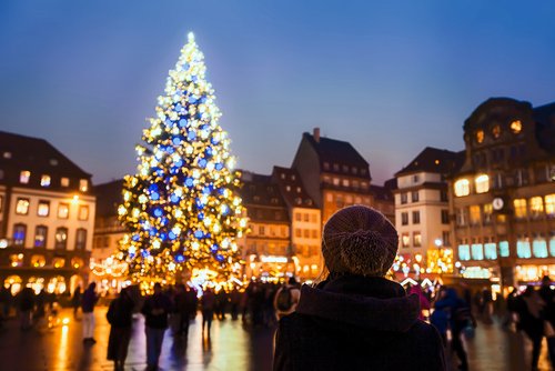 Eine Frau mit Mütze von hinten vor einem Weihnachtsmarkt in Straßburg bei Nacht, mit festlich beleuchteten Ständen zwischen historischen Gebäuden und einem großen, beleuchteten Weihnachtsbaum.