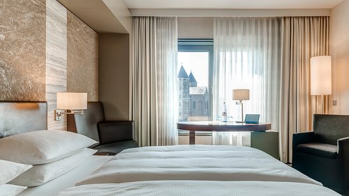 Ein modernes Hotelzimmer des Hotel Dorint in Köln mit Doppelbett, einem Sessel, einer Sitzbank, einem runden Tisch vor einem Fenster mit Blick auf eine Kirche mit zwei Türmen.