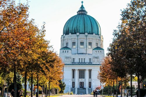 Eine Allee mit herbstlichen Bäumen führt zu einer Kirche mit Dachkuppel und Kreuz auf dem Wiener Zentralfriedhof.