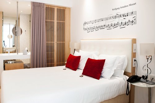 Hotelzimmer mit großem Bett, weißen Bettwäsche, zwei roten Kissen, Wanddekoration mit Noten von Wolfgang Amadeus Mozart und Nachttisch mit Telefon und Lampe in Form eines Violinschlüssels.
