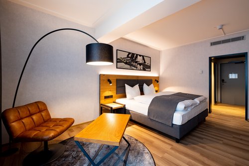 Doppelzimmer im MK Hotel Passau mit Doppelbett, braunem Ledersessel, Bogenlampe und Holztisch