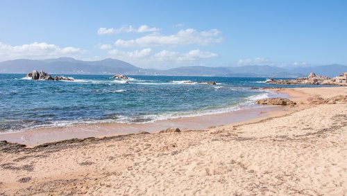 Strand von Port-Saint-Louis mit feinem Sand, ruhigem Wasser unter blauem Himmel. Im Hintergrund sind Berge und eine Stadt