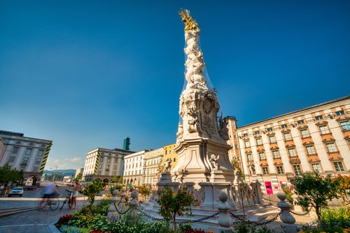 Blick auf einen öffentlichen Platz in Linz unter blauem Himmel. Im Vordergrund steht eine beige historische Statue, im Hintergrund sind bunte Wohnhäuser.
