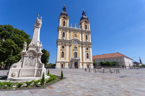 Barocke Kathedrale in Kalocsa, Ungarn, mit zwei Türmen und einer Kuppel, umgeben von Bäumen und einem blauen Himmel im Hintergrund.