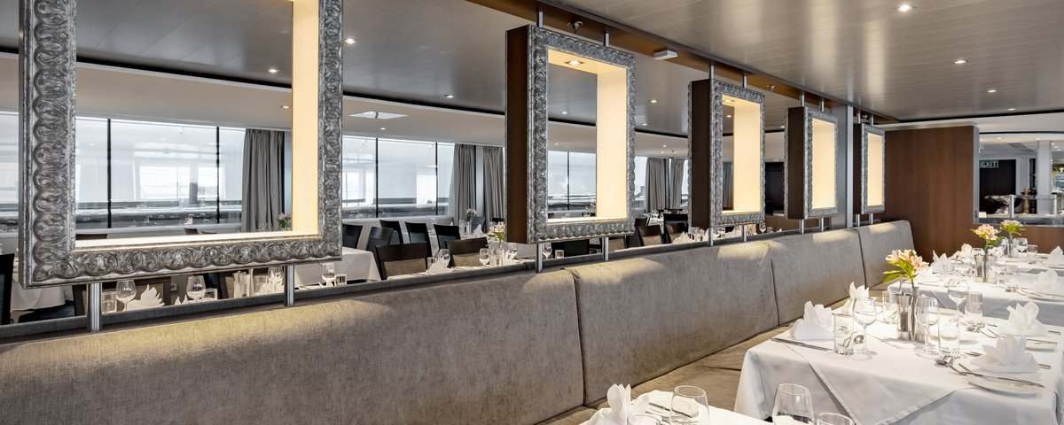 Stilvoll eingedeckter Restaurantbereich der A-ROSA ALEA mit Fenstern, Blumendeko und gedämpfter Beleuchtung.