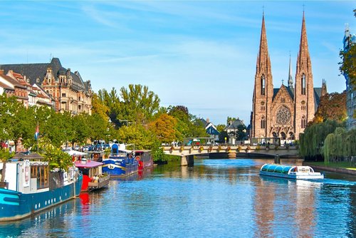 Straßburger Altstadt mit Fachwerkhäusern und Kanälen und einer Kirche, umgeben von grünen Bäumen, unter blauem Himmel.