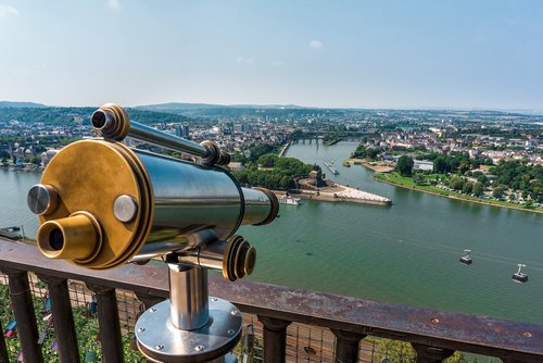 Ein Fernrohr auf einem Aussichtspunkt mit Blick auf die Stadt Koblenz und das Deutsche Eck, eine Hochseilbahn fährt über den Fluss bei klarem Himmel.