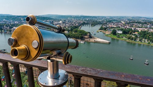 Ein Fernrohr auf einem Aussichtspunkt mit Blick auf die Stadt Koblenz und das Deutsche Eck, eine Hochseilbahn fährt über den Fluss bei klarem Himmel.